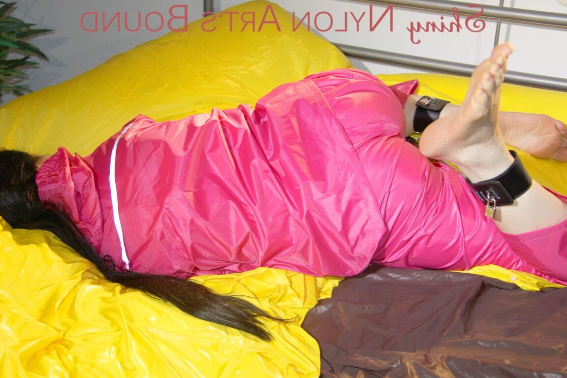 16P ルーシーはセクシーなピンクの光沢のあるナイロンの雨を着てベッドの上でバーで縛られて猿ぐつわをされています