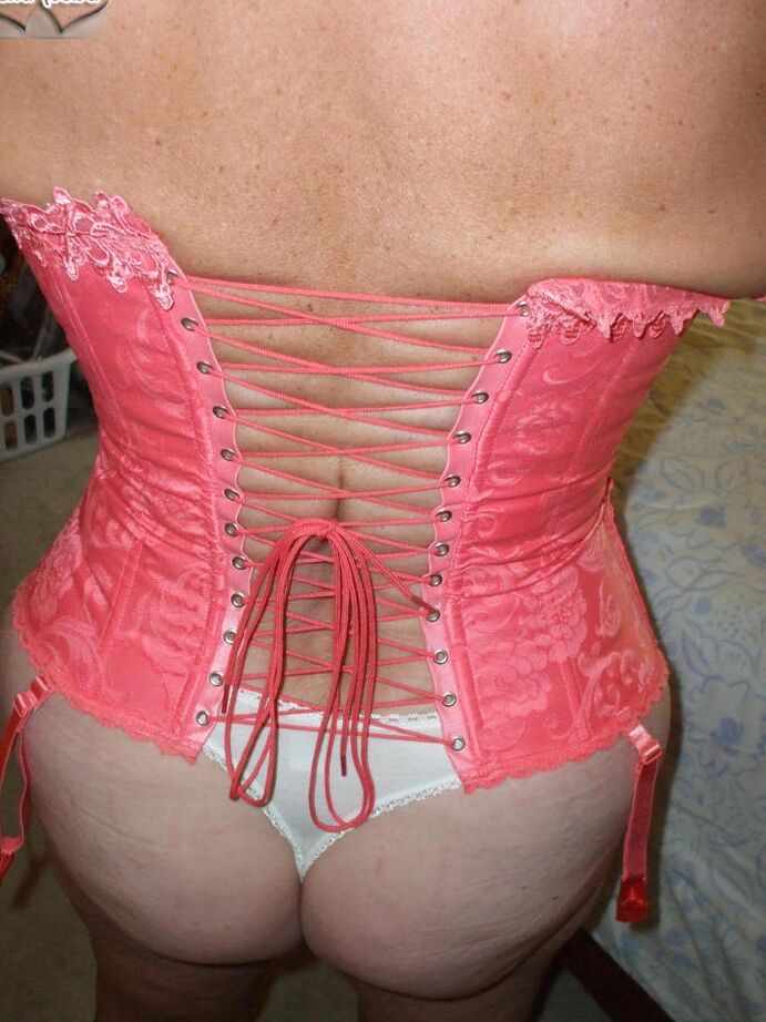 19P Busty Bliss, amateur mature, présente un corset lacé et un string pieds nus.