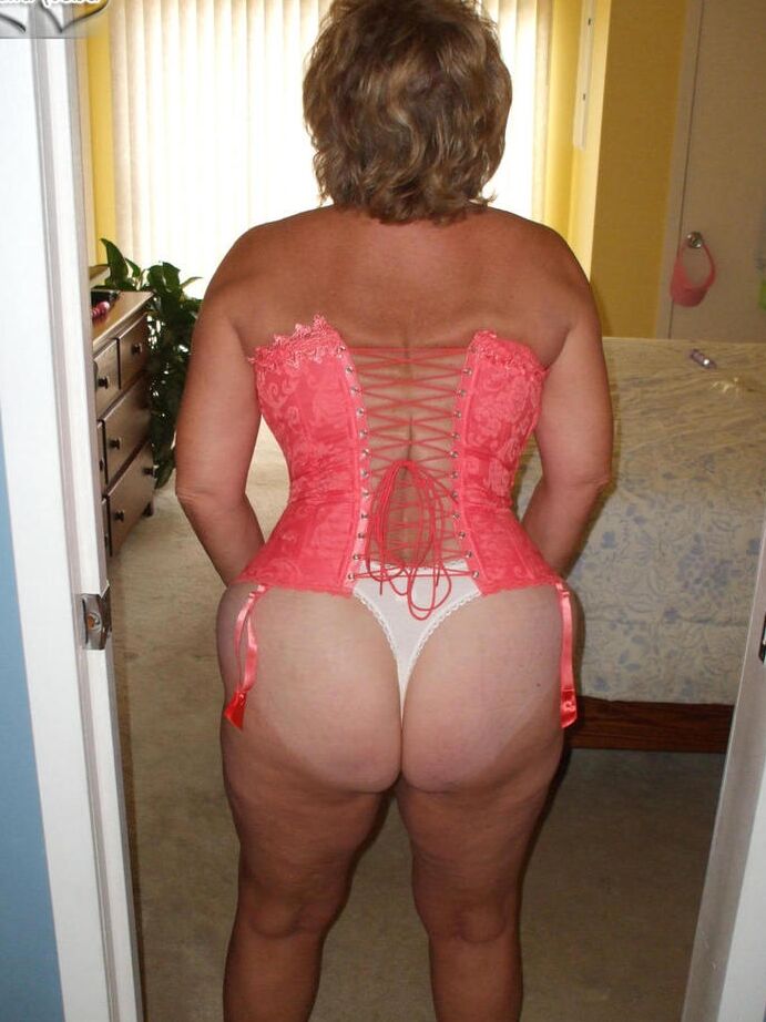 19P Busty Bliss, amateur mature, présente un corset lacé et un string pieds nus.
