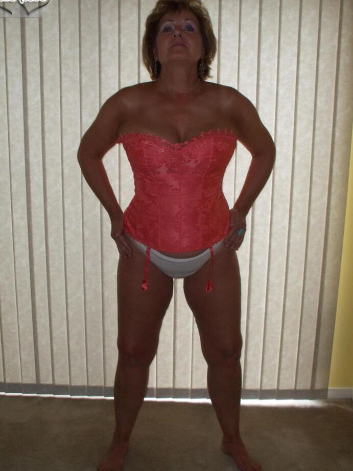 19P Busty Bliss, amateur mature, présente un corset lacé et un string pieds nus.