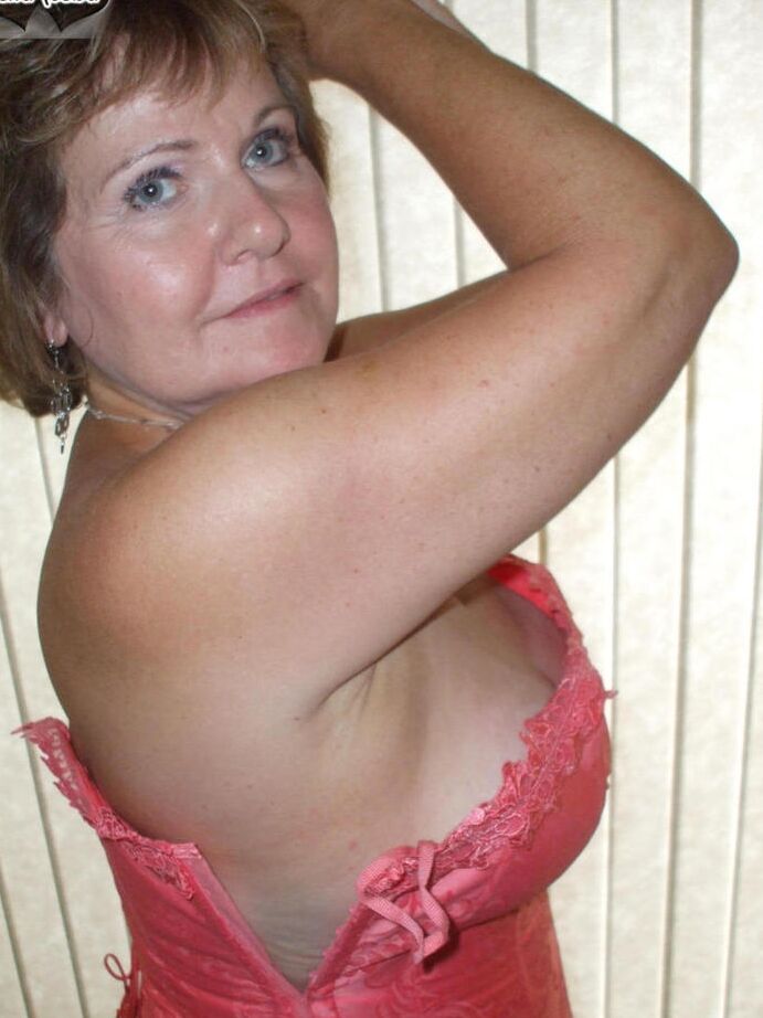 19P Busty Bliss, amateur mature, présente un corset lacé et un string pieds nus.