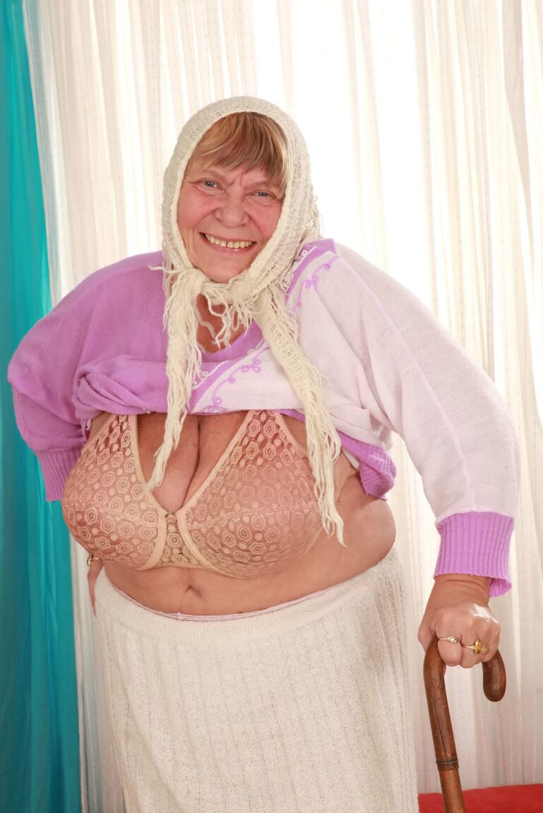 15P Old granny Sara G in skirt & boots sheds sweater to show huge tits in bra