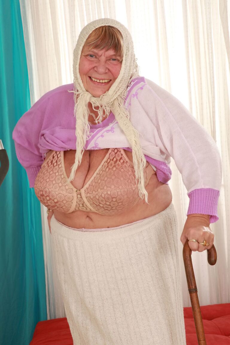 15P Old granny Sara G in skirt & boots sheds sweater to show huge tits in bra