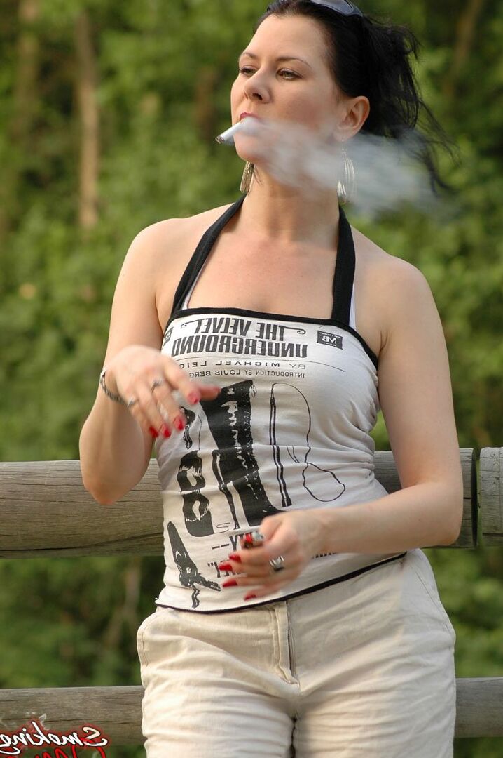 15P Solo girl smokes a cigarette up against a rail fence while fully clothed