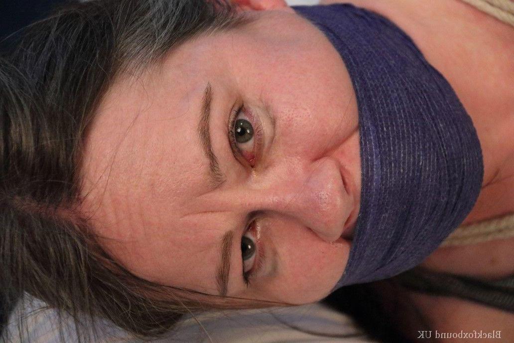 16P Kaukasische Frau liegt geknebelt und gefesselt mit dem Gesicht nach unten auf einem Bett
