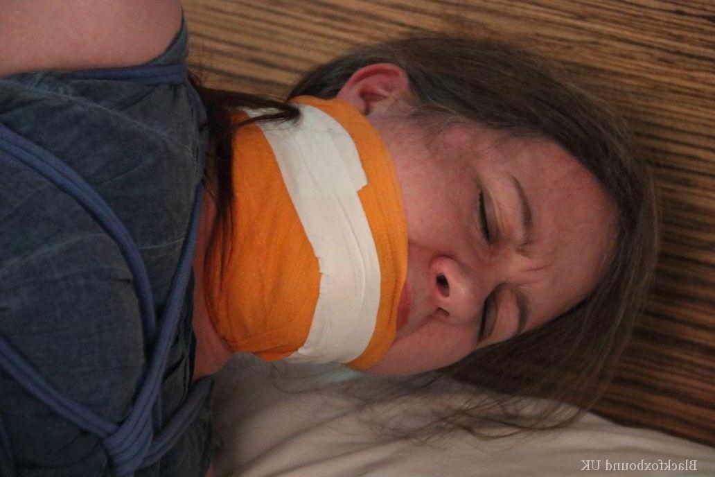 16P Kaukasische Frau liegt geknebelt und gefesselt mit dem Gesicht nach unten auf einem Bett