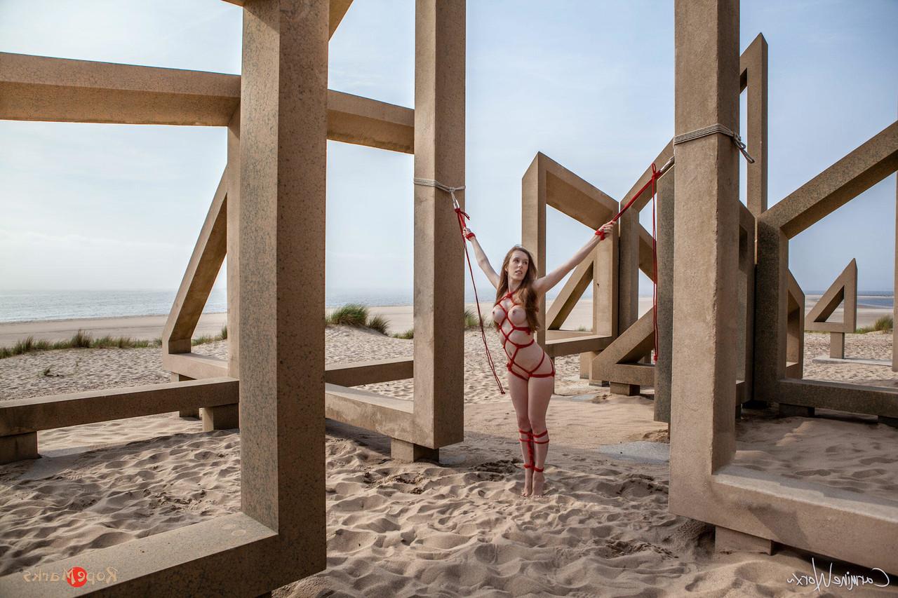 16P White girl Carmine Worx is tied with ropes to construction beams on a beach