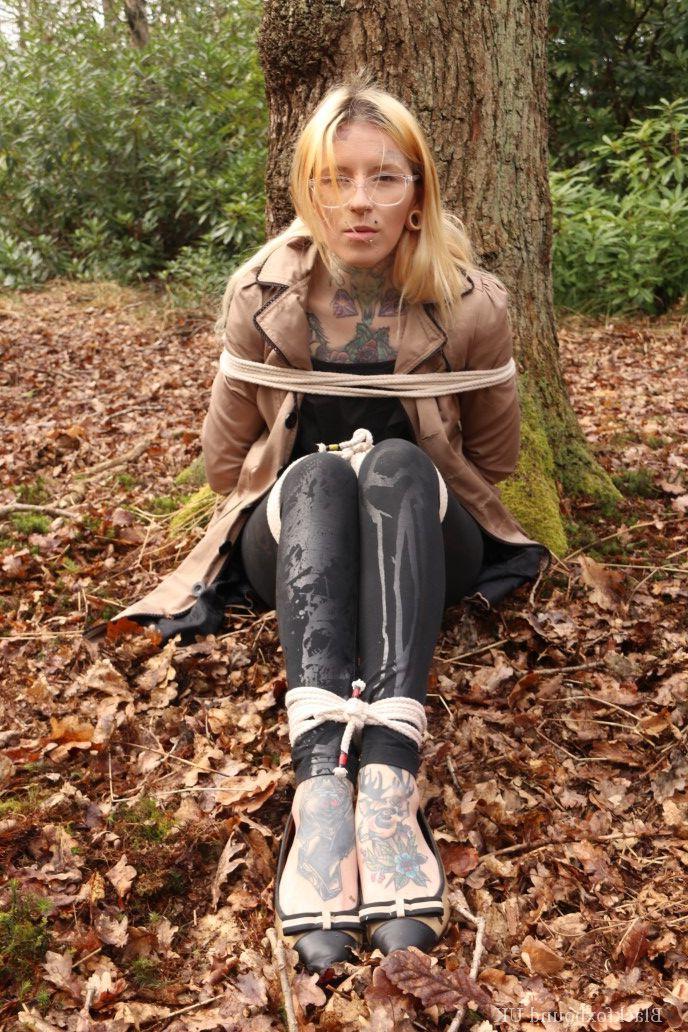 16P Eine Frau mit Brille ist mit Seilen an einen Baum im Wald gefesselt und trägt Kleidung