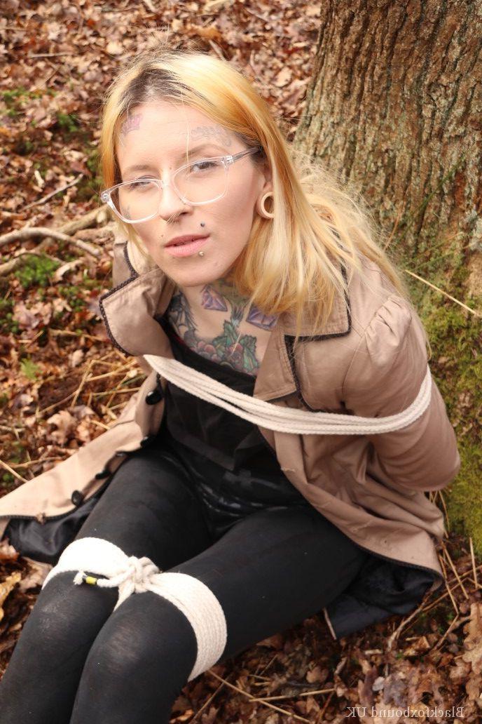 16P Eine Frau mit Brille ist mit Seilen an einen Baum im Wald gefesselt und trägt Kleidung