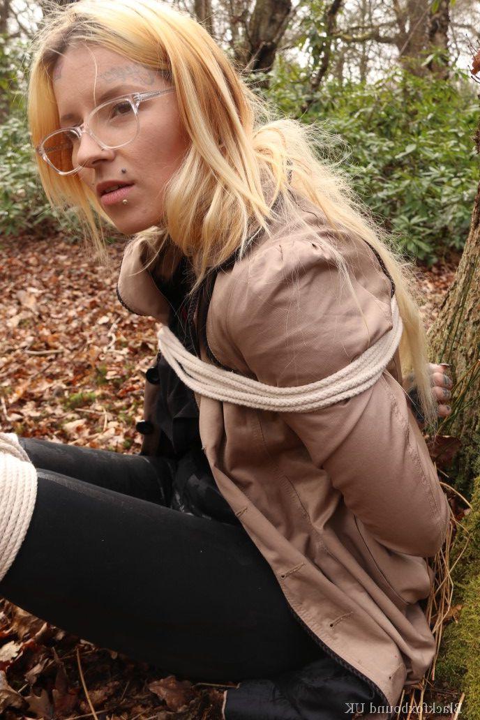 16P Eine Frau mit Brille ist mit Seilen an einen Baum im Wald gefesselt und trägt Kleidung