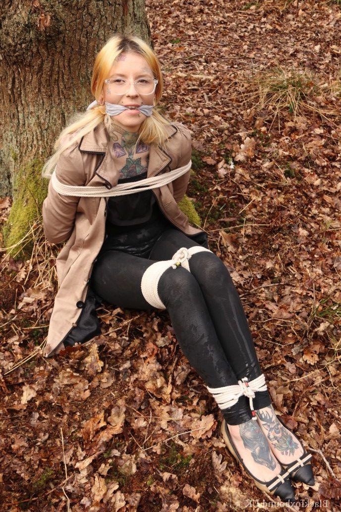 16P Eine Frau mit Brille ist mit Seilen an einen Baum im Wald gefesselt und trägt Kleidung
