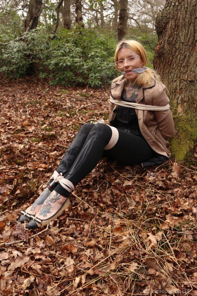 16P Eine Frau mit Brille ist mit Seilen an einen Baum im Wald gefesselt und trägt Kleidung