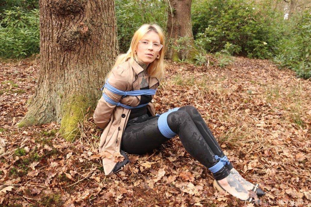 16P Eine Frau mit Brille ist mit Seilen an einen Baum im Wald gefesselt und trägt Kleidung