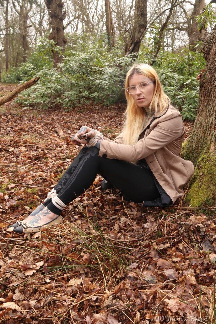 16P Eine Frau mit Brille ist mit Seilen an einen Baum im Wald gefesselt und trägt Kleidung