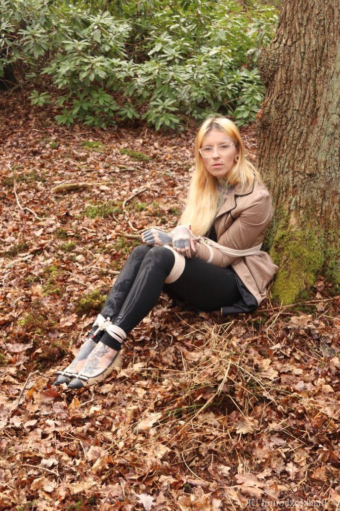 16P Eine Frau mit Brille ist mit Seilen an einen Baum im Wald gefesselt und trägt Kleidung