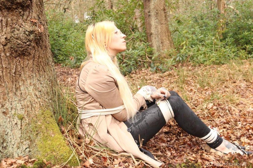 16P Eine Frau mit Brille ist mit Seilen an einen Baum im Wald gefesselt und trägt Kleidung