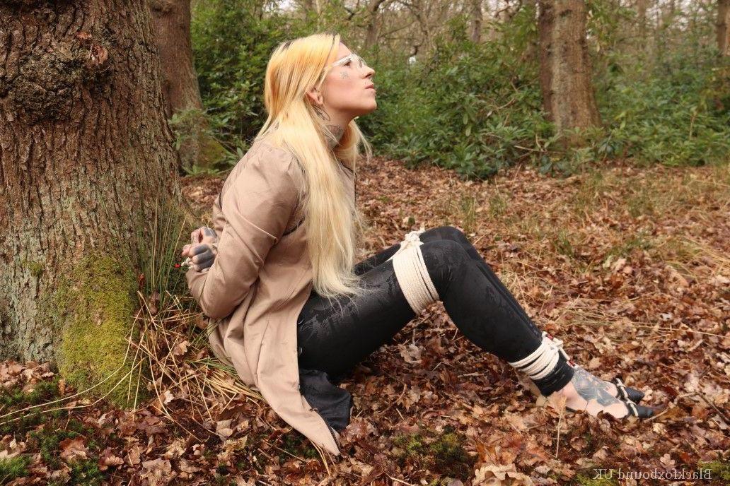 16P Eine Frau mit Brille ist mit Seilen an einen Baum im Wald gefesselt und trägt Kleidung