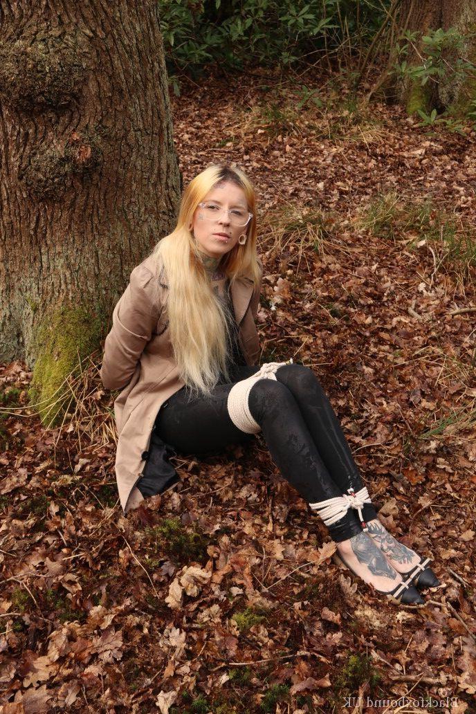 16P Eine Frau mit Brille ist mit Seilen an einen Baum im Wald gefesselt und trägt Kleidung