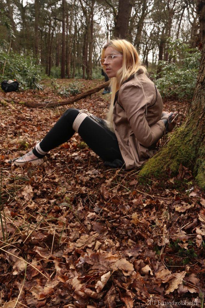 16P Eine Frau mit Brille ist mit Seilen an einen Baum im Wald gefesselt und trägt Kleidung