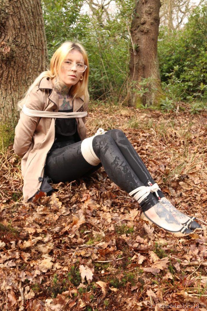 16P Eine Frau mit Brille ist mit Seilen an einen Baum im Wald gefesselt und trägt Kleidung