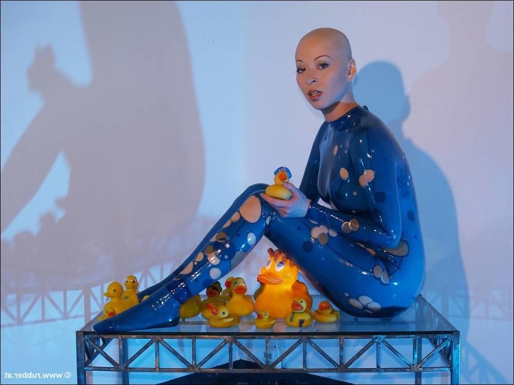 16P Bald female models rubber attire during a solo engagement