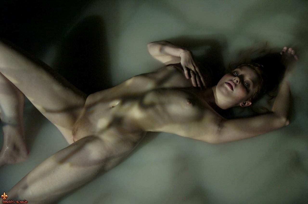 15P Jeny, la belle rousse, est allongée nue dans une eau peu profonde, sous un faible éclairage.