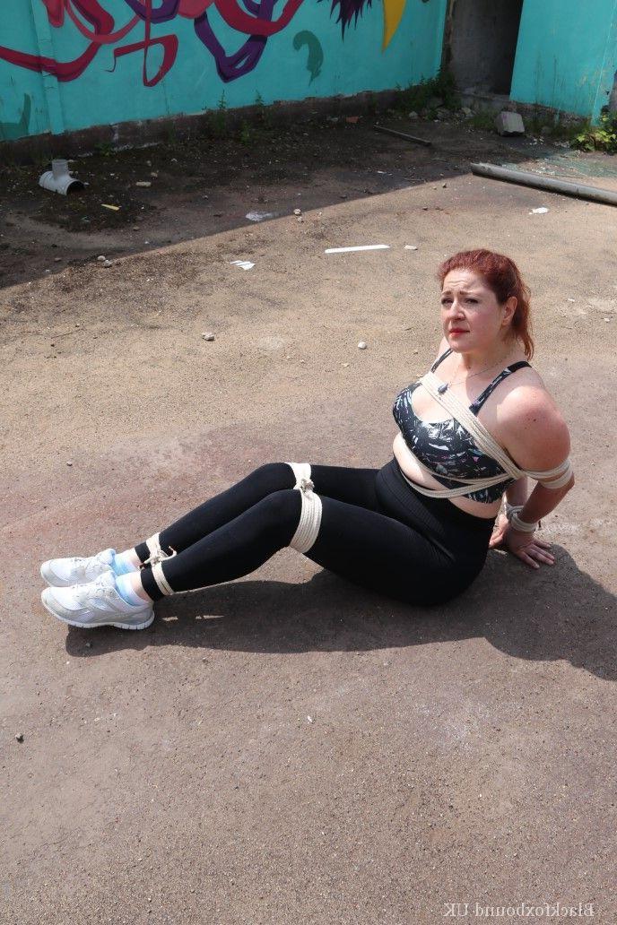 16P Redheaded girl Mada Rose is left on a paved lot while tied up