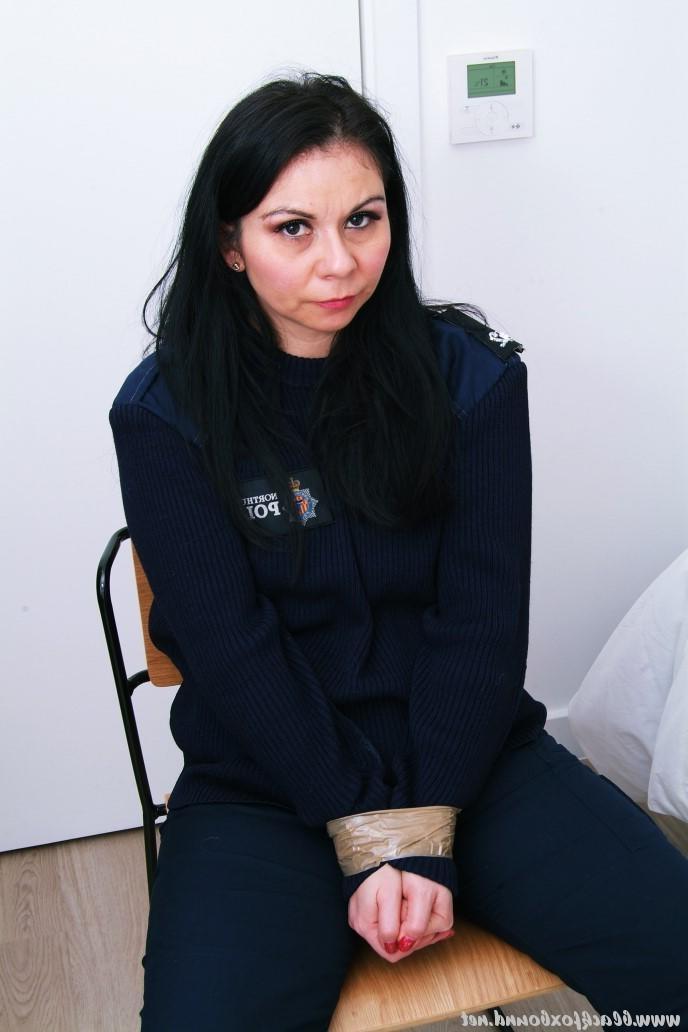 16P Mujer policía de cabello oscuro se encuentra atada a una silla con su uniforme