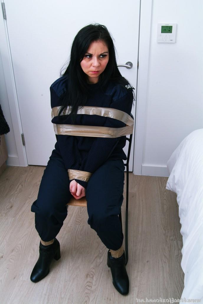 16P Mujer policía de cabello oscuro se encuentra atada a una silla con su uniforme
