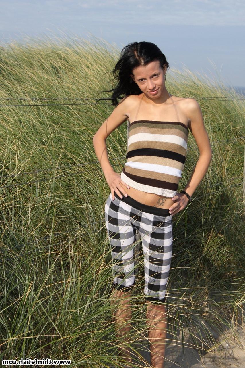 16P Une brune mince tient son cul nu pieds nus sur une plage de sable